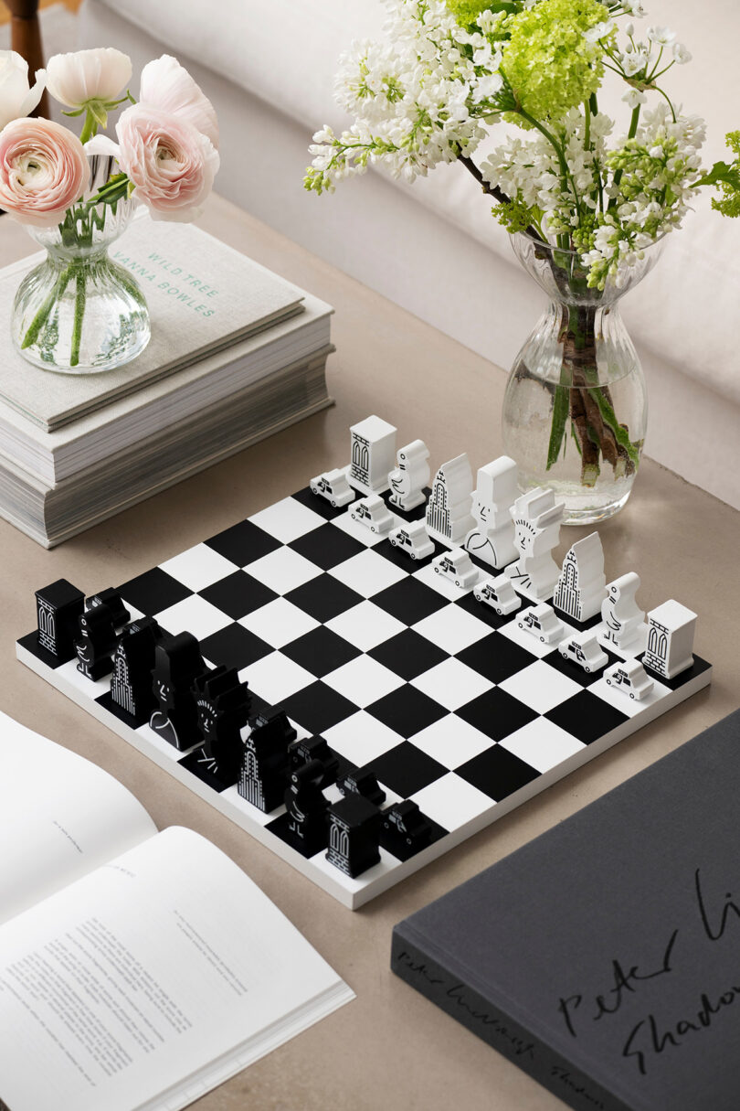 A black and white chess set with architectural pieces on a table, surrounded by stacked books and vases with flowers.