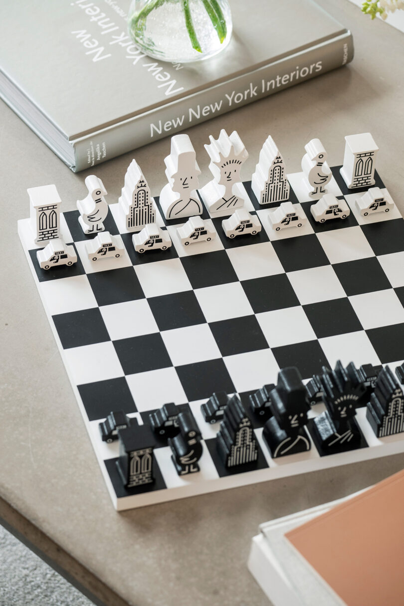 Chess set with pieces designed as stylized city buildings and figures. The board is black and white, positioned on a table with books in the background.