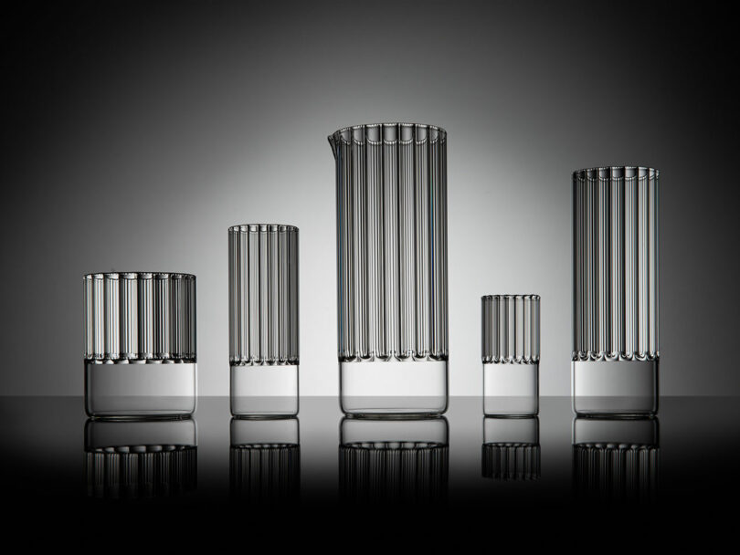 A set of five transparent, vertically striped glass vessels by Felicia Ferrone stand gracefully in a row, their varying heights creating a stunning silhouette against a gray background.