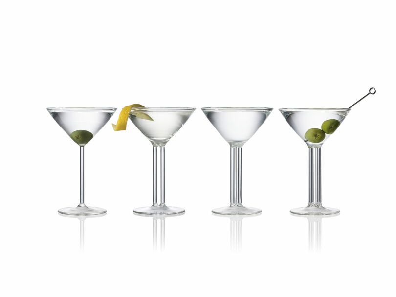 Four martini glasses in a row, echoing the sophisticated design of Felicia Ferrone. The first has an olive, the second a lemon twist, the third is plain, and the fourth boasts olives on a skewer.