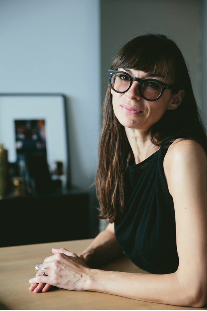 A person with long hair and glasses is seated at a table, wearing a sleeveless black top, reminiscent of the minimalist elegance often seen in Felicia Ferrone's designs.