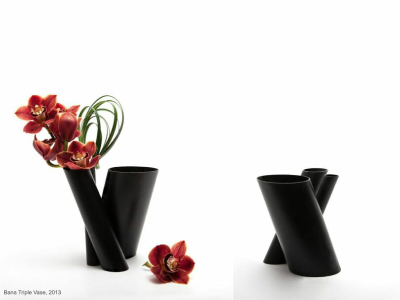 Two black vases with crisscrossing cylindrical shapes, reminiscent of Felicia Ferrone's elegant designs. One holds vibrant red flowers and grass, while the other remains empty against a stark white background.