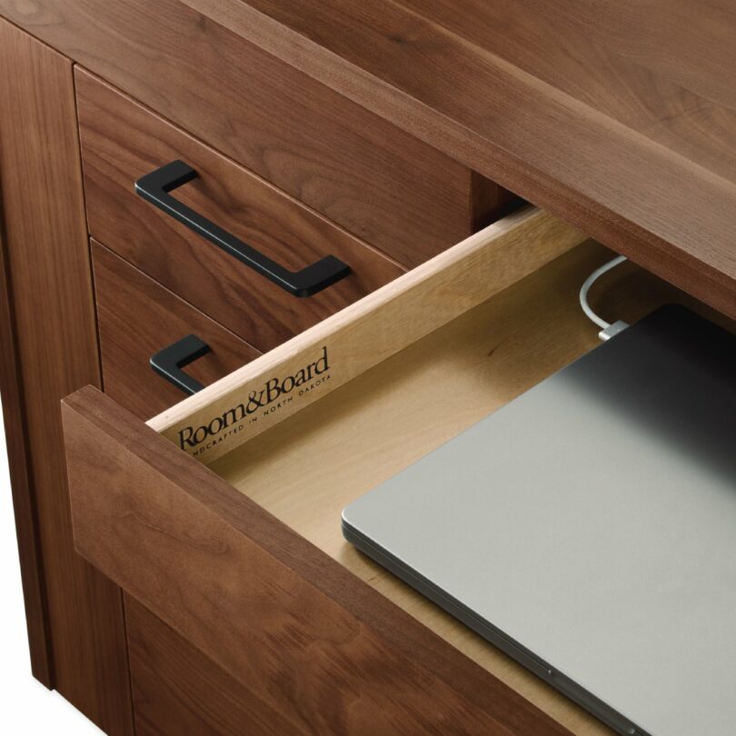 Wooden desk with a partially open drawer, revealing a silver laptop inside. Black handles are visible on the drawers, and branding reads "Room & Board.