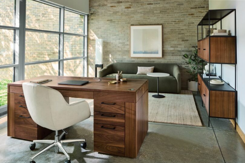 Modern office with wooden desk, white swivel chair, gray sofa, and shelves. A large window lets in natural light. A framed picture hangs on the brick wall.