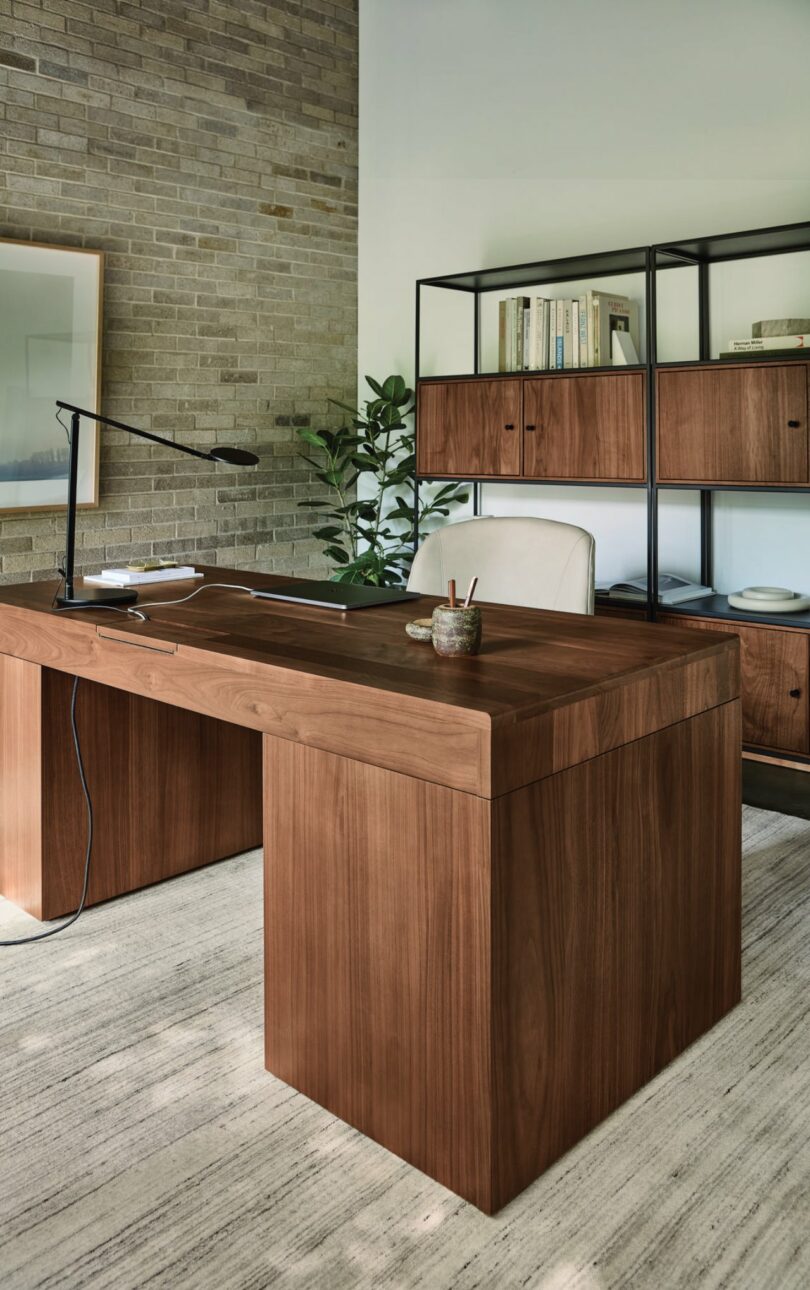 Minimalist office with a wooden desk, modern lamp, chair, and shelving unit. Brick wall backdrop with a plant and artwork. Neutral tones and natural lighting.