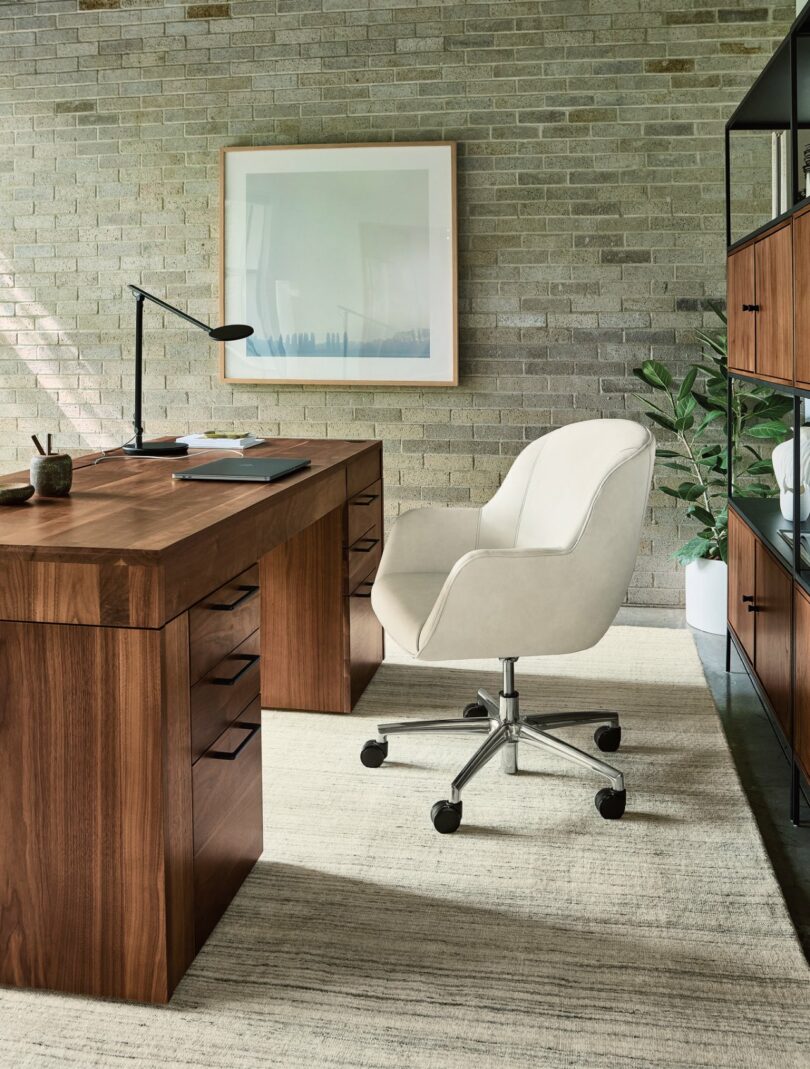 Modern office with a wooden desk, white swivel chair, lamp, and shelves. Framed picture on brick wall and green plant in the corner. Neutral tones throughout the room.