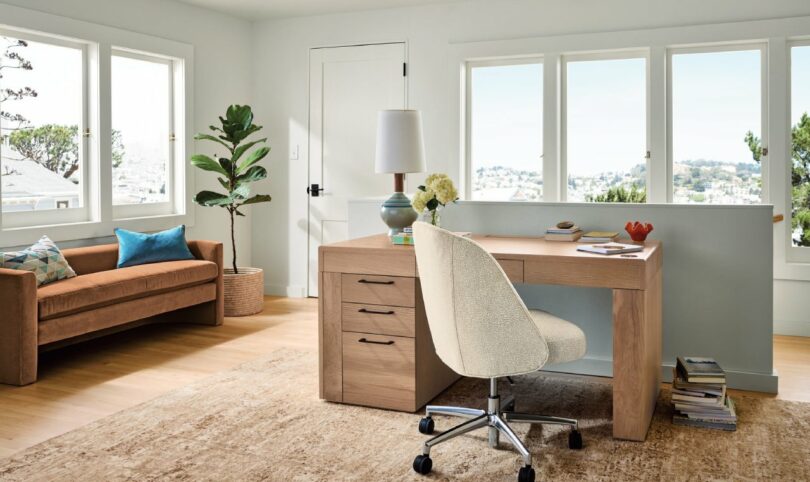 A home office with a wooden desk, beige swivel chair, and brown sofa, featuring large windows, a potted plant, and a lamp on the desk. A rug covers the wooden floor.