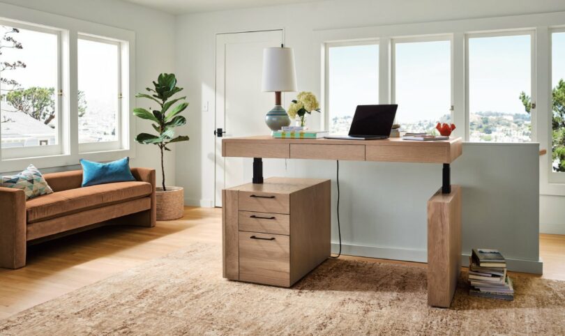 Modern office with a standing desk, laptop, and lamp, near a large window. A brown sofa with colorful cushions is on the left, and a potted plant is in the corner. Neutral-colored rug on floor.