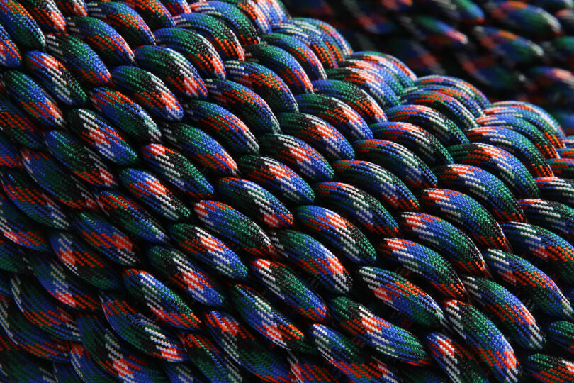 Close-up of a tightly woven paracord, reminiscent of SHORE-inspired footwear, showcasing a pattern of blue, green, red, and black strands that create a textured, braided appearance.