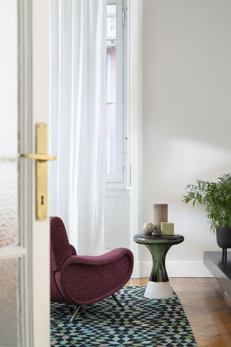 Room interior with a red armchair, geometric rug, and small green table with a candle and books. Nearby is a plant on a low cabinet, and sunlight streams through a white curtained window.