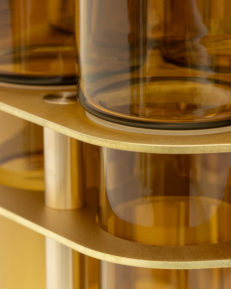 Close-up of transparent amber cylinders arranged on a tiered, golden-hued structure with vertical supports. The cylinders reflect light, creating a warm and reflective appearance.