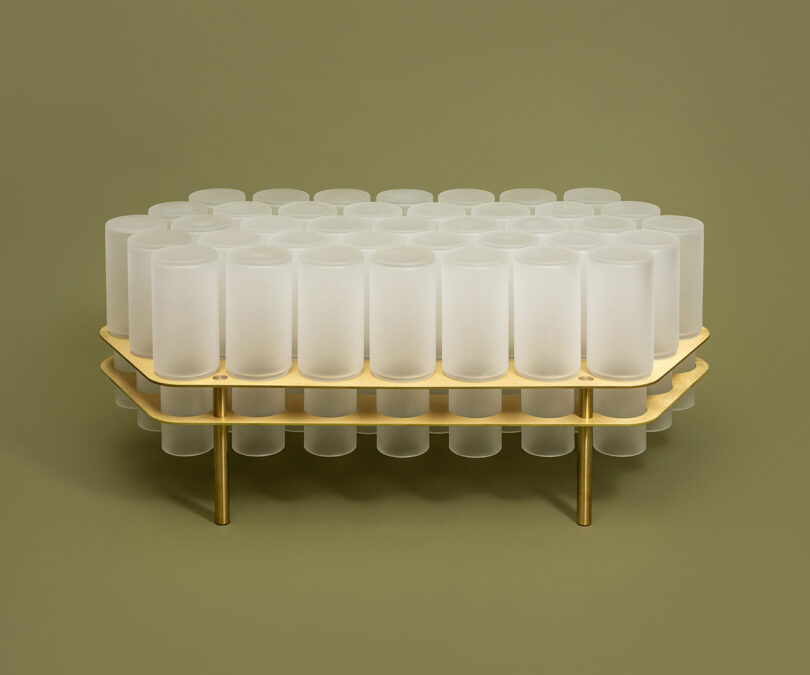 A modern table with two gold-colored metal layers, each supporting rows of white cylindrical containers, set against a plain olive-green background.