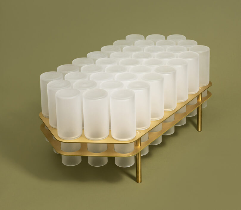 A modern table with a gold frame featuring multiple translucent, cylindrical stubs on top, set against a muted green background.