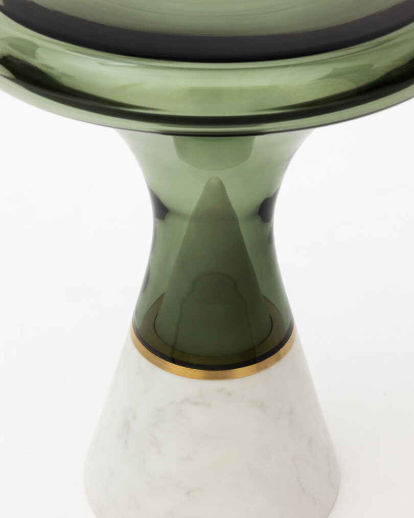 A green glass object with a wide top, narrowing to a central stem, set on a white, conical marble base with a gold band.