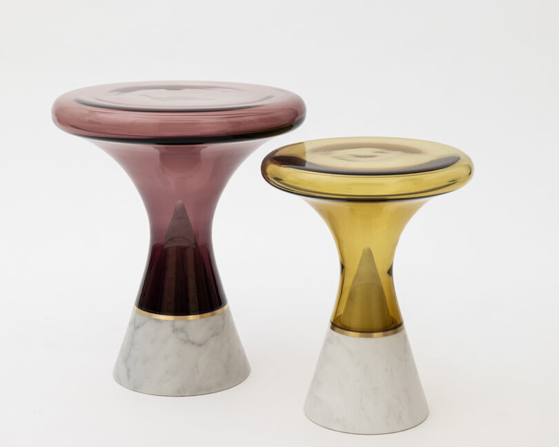 Two glass-top tables with conical marble bases. One table is taller with a purple top, and the other is shorter with a yellow top, both against a plain white background.