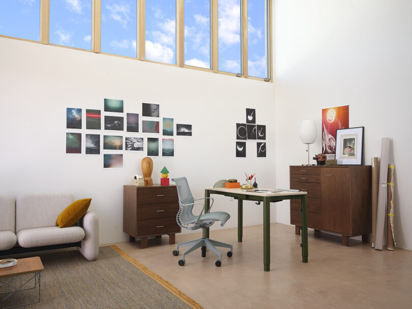 Modern home office with a green desk, mesh chair, and wall art. A rug lies on the floor beside a sofa. Wooden cabinets hold decor items and a lamp. Natural light enters through a high window.