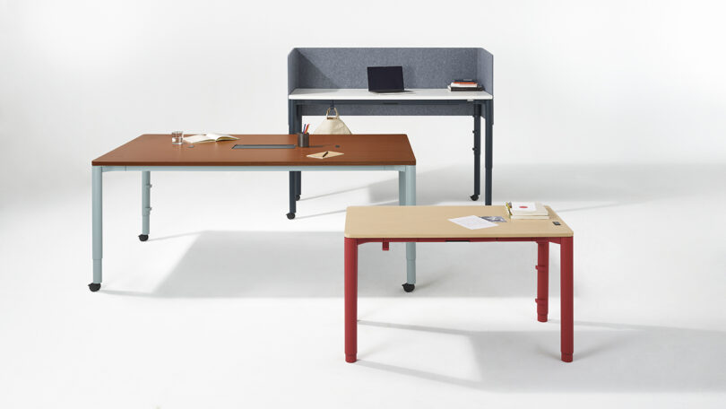 Three modern desks with adjustable heights: a large brown desk with items, a gray desk with a laptop and privacy screen, and a smaller beige desk with red legs, on a white background.