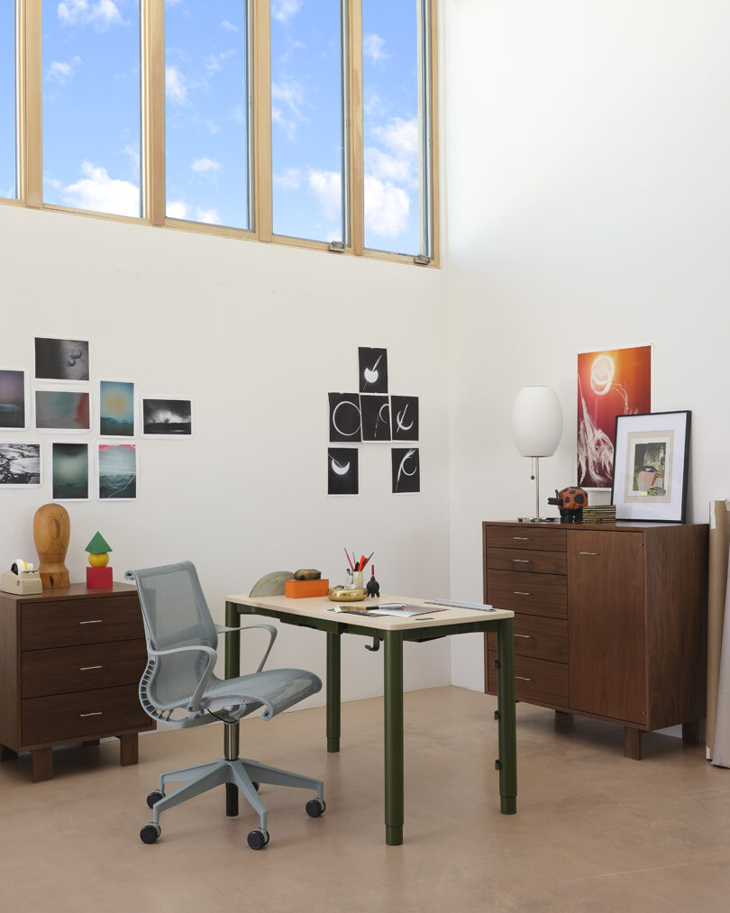 Modern office space with a desk, ergonomic chair, two wooden cabinets, and framed art on white walls. Large windows reveal a blue sky.