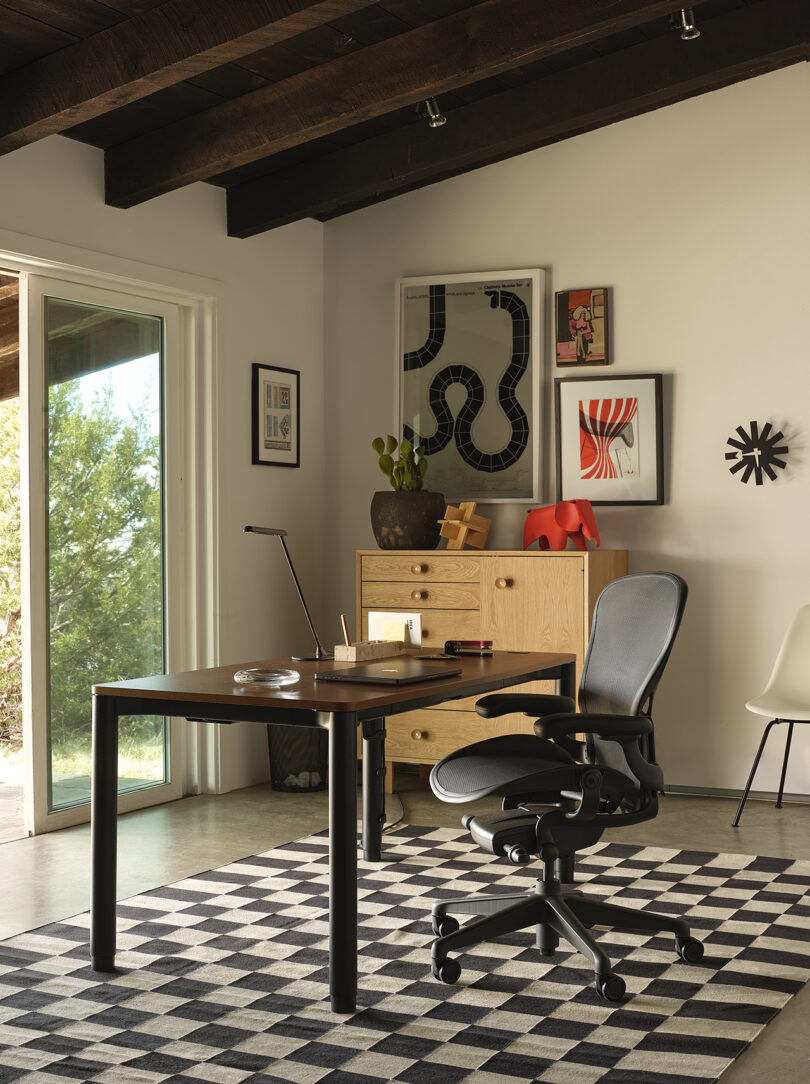 Modern home office with a black desk, ergonomic chair, and decor including framed art and a checkerboard rug. Large window offers natural light and view of trees.