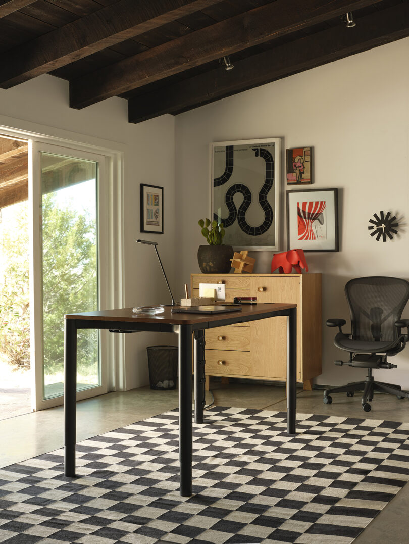 Home office with a modern desk and chair on a checkered rug. Art and decor on white walls, a wooden cabinet with a plant, and a large window overlooking greenery.