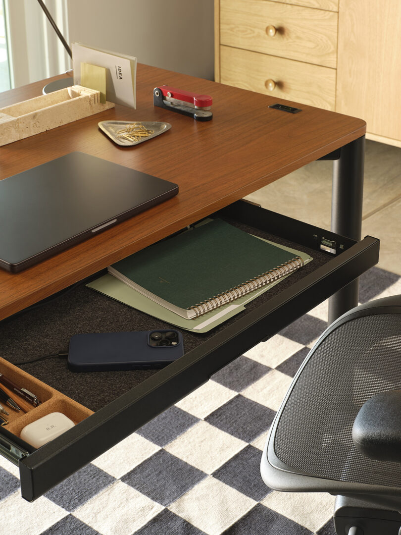 Wooden desk with a drawer open, containing notebooks and a smartphone. Laptop, business cards, and a utility knife are on top; chair and checkered rug visible.