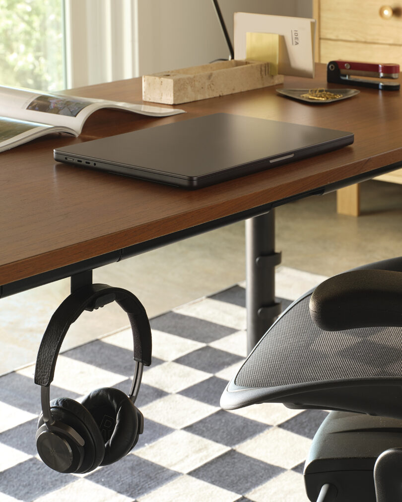 Wooden desk with a closed laptop, an open magazine, a pencil, headphones hanging off the side, and a checkered rug underneath.