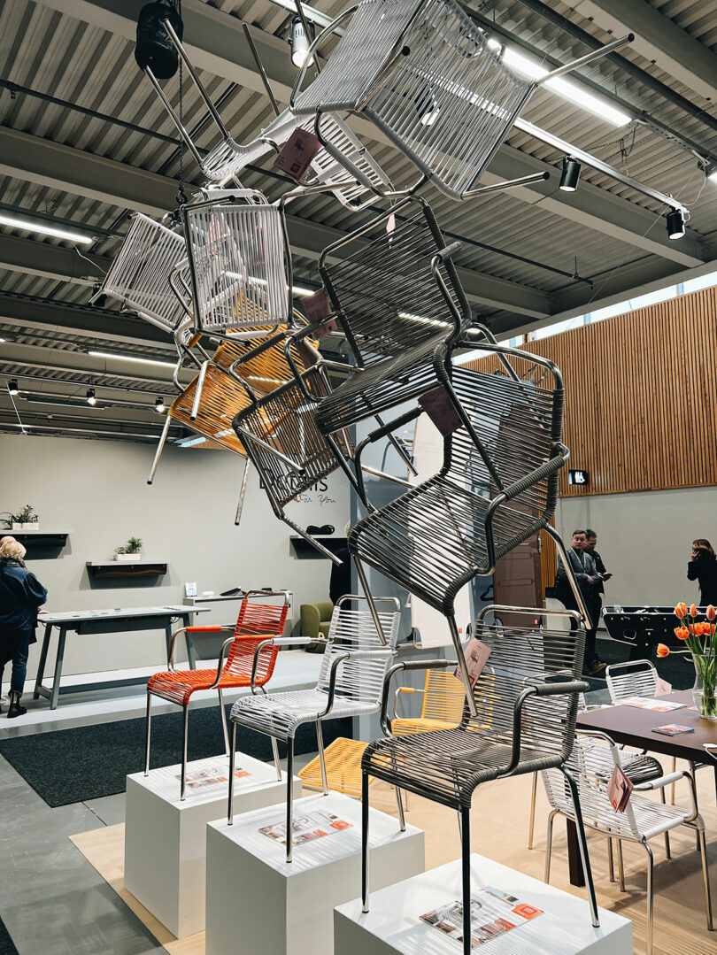 Chairs balanced in a vertical stack on white pedestals create an artistic display reminiscent of the Stockholm Furniture Fair, with curious onlookers admiring the innovative designs in the showroom's background.