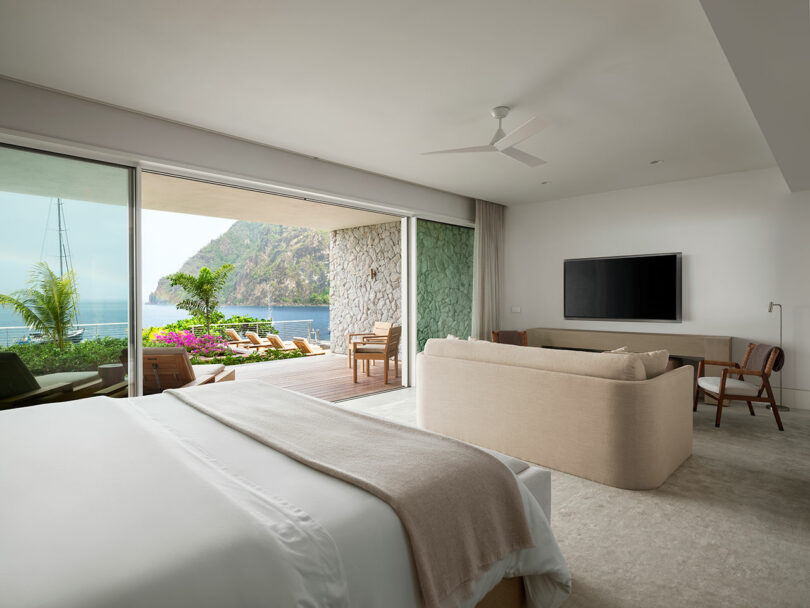 Luxury hotel room with beige bed and sofa, TV, and an open sliding glass door leading to a balcony with ocean and cliff views. Outdoor seating and plants are visible.