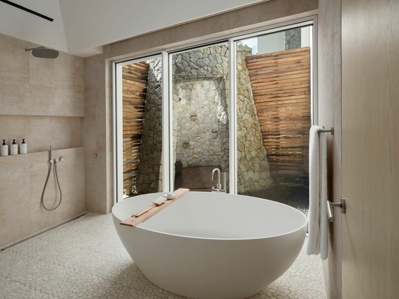 Modern bathroom with freestanding bathtub, large windows revealing a stone and wood privacy wall. Pebble flooring and a wall-mounted shower are visible.