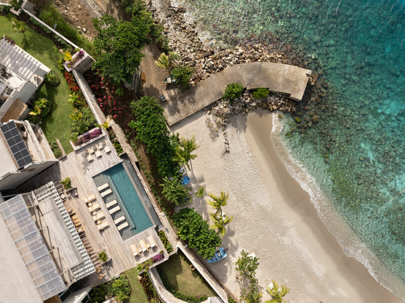 Aerial view of a beach resort featuring an infinity pool with lounge chairs, adjacent to a sandy beach and rocky shoreline, bordered by lush greenery and clear turquoise water.