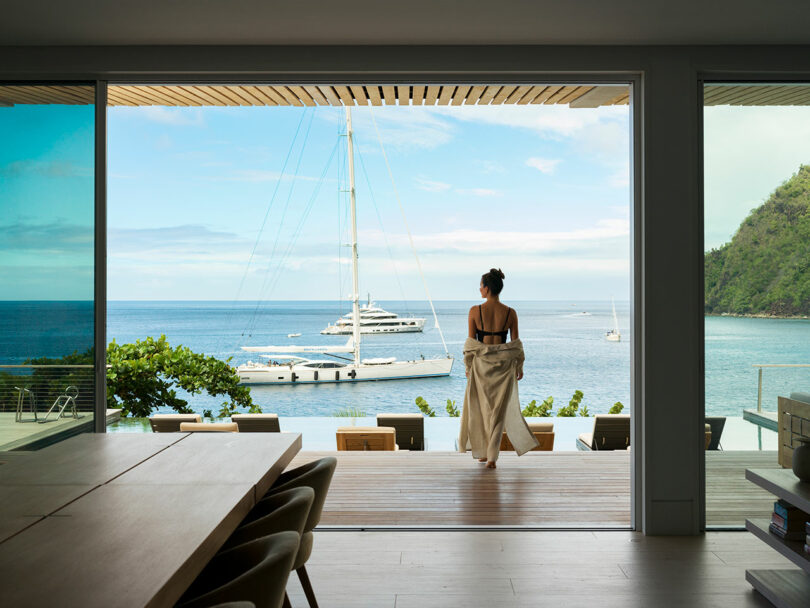 Person in swimwear stands on a patio overlooking a bay with yachts and a lush hillside under a clear sky.