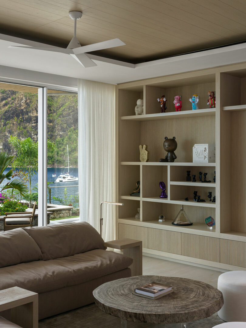 Modern living room with a beige sofa, round wooden coffee table, ceiling fan, and shelf displaying decorative items. Large window offers a view of a patio, lush greenery, and a marina with boats.