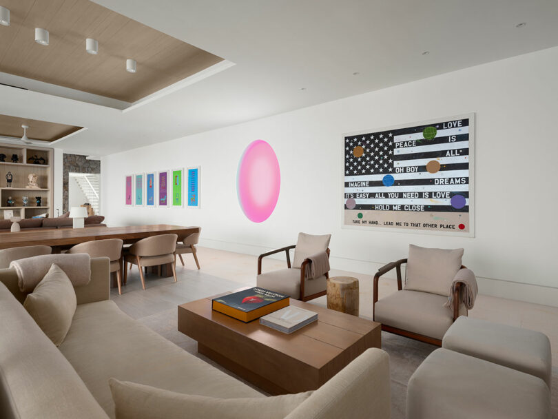 Minimalist living room with neutral furniture, wooden coffee table, and colorful modern art on white walls. Ceiling features recessed lighting, room includes dining area with a long table.