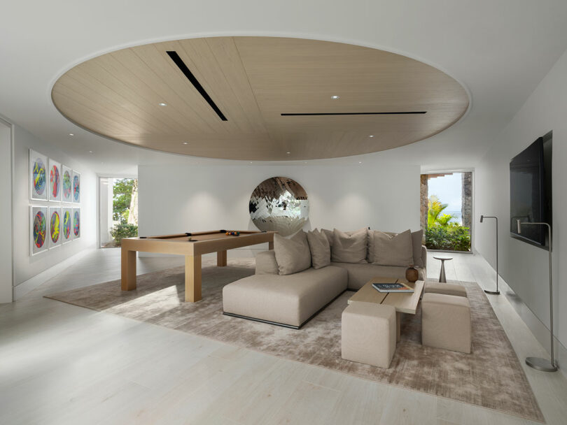 Modern living room with a large beige sectional sofa, a round wooden ceiling feature, a pool table, colorful wall art, and a view of greenery outside.