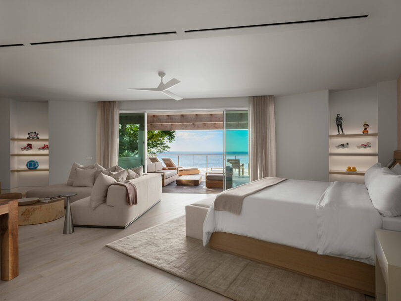 Modern bedroom with a white bed, seating area, and open glass doors leading to a balcony with ocean view. Shelves with decor items are on the walls, and a ceiling fan is above.