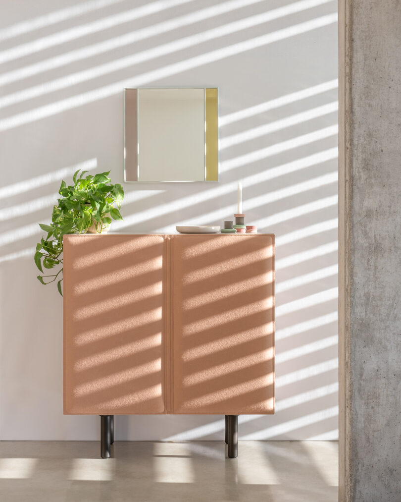 Tan cabinet against a white wall with diagonal shadows. A plant and candle sit on top, with a square mirror above.