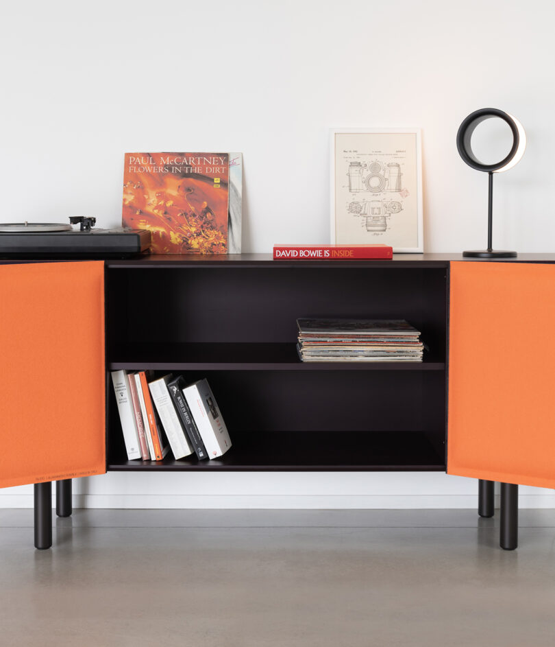 A black console with orange doors displays records and books. Paul McCartney album and framed print on top, with a lamp. A David Bowie book on the shelf below.