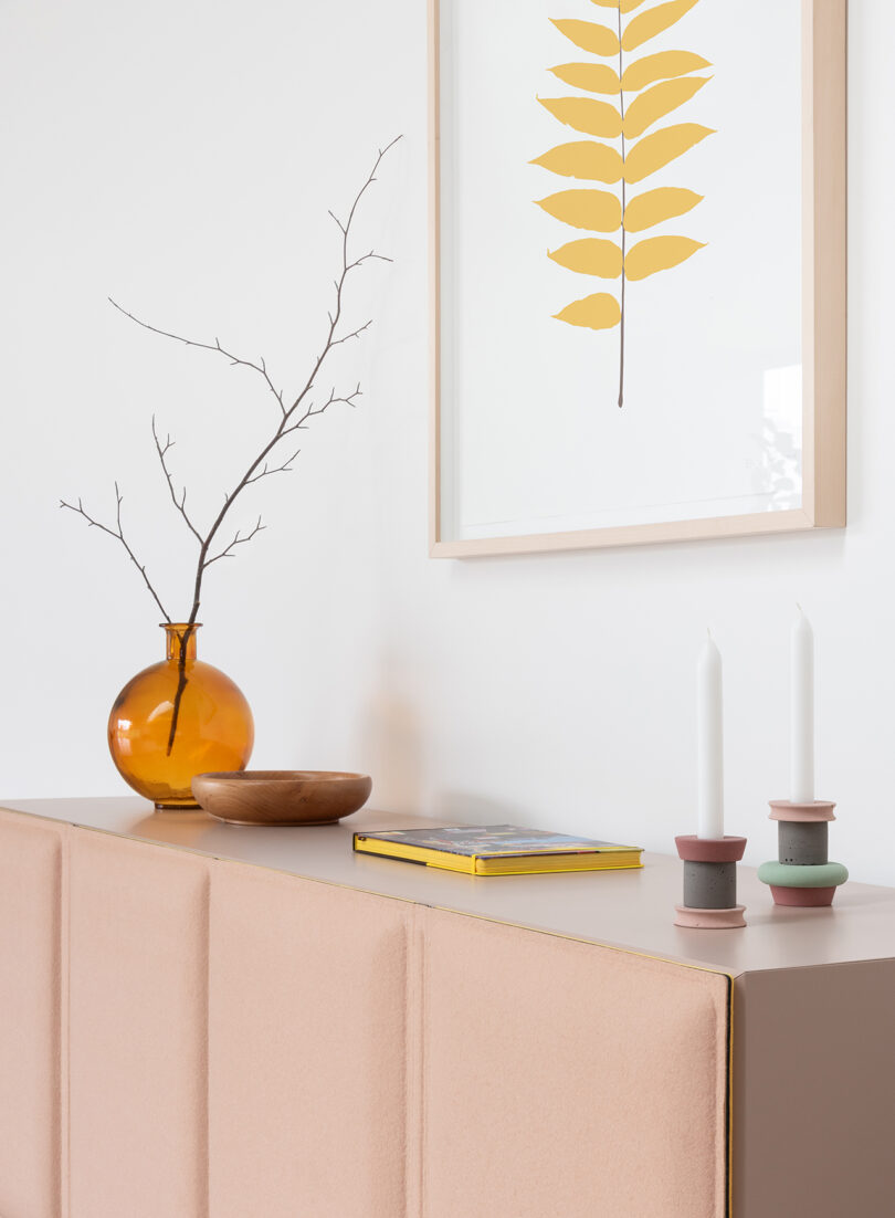 Minimalist interior with a beige cabinet, an amber vase holding twigs, a wooden bowl, a book, candle holders, and a framed botanical print on the wall.