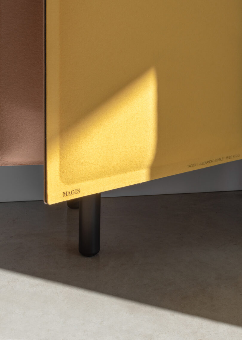 A yellow panel with the word "Magis" printed on it casts a shadow on the floor. The panel stands on a small black cylindrical leg next to a wall.