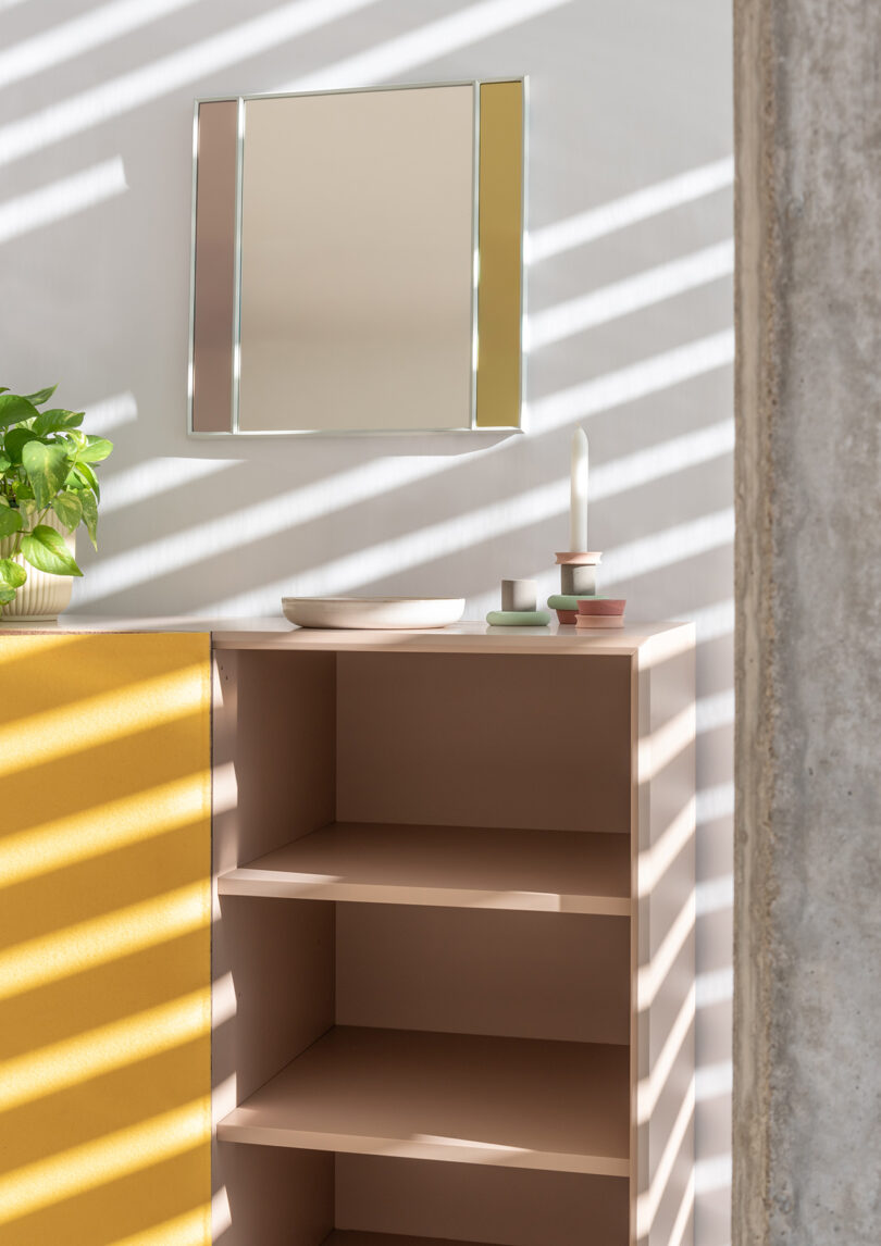 A beige shelf with a white candle, small trays, and a plant beside it. A mirror hangs above, and diagonal shadows from blinds fall across the scene.
