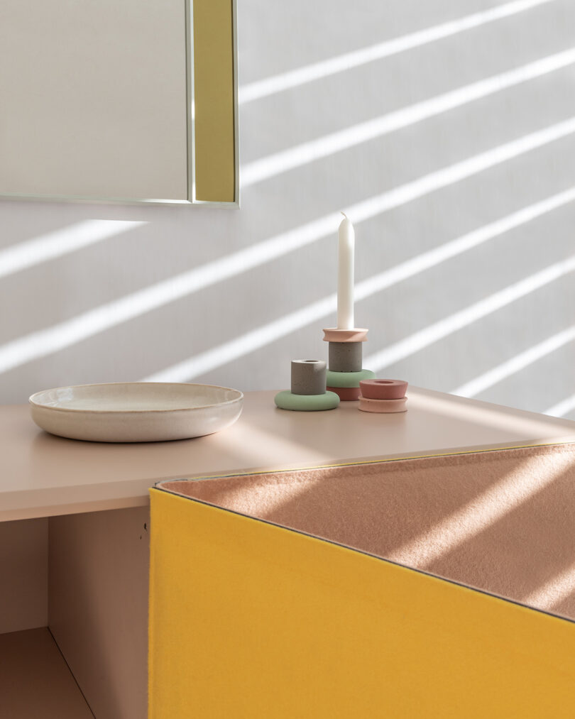 Minimalist interior with a tabletop displaying a white candle, small ceramic dishes, and a bowl. Soft light from window blinds casts diagonal shadows on the wall.