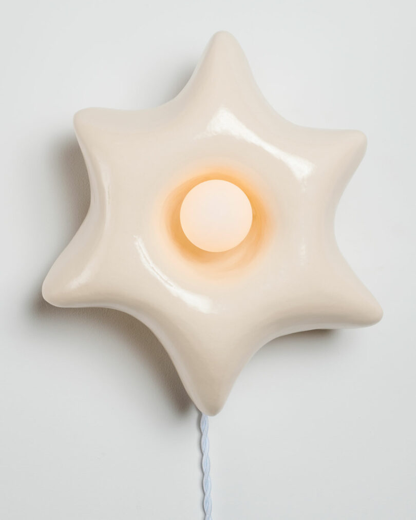 A star-shaped, cream-colored lamp with a glowing central bulb and a visible cord hangs on a plain white wall.