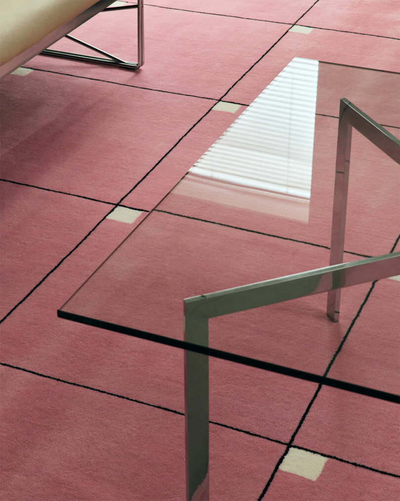 Glass coffee table with metal frame on a pink grid-patterned carpet. Part of a modern beige sofa visible in the corner.