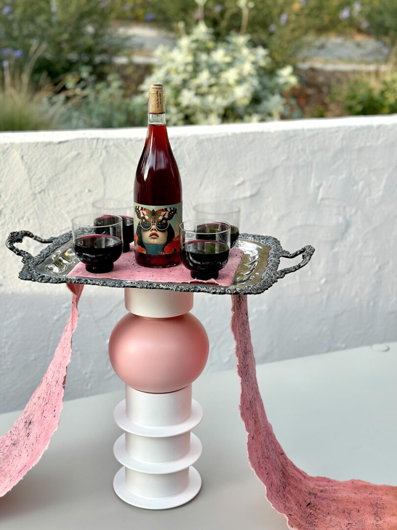 A stylized wine display featuring a bottle and glasses on an ornate tray, placed atop a sculptural pedestal with pink paper draping over the sides.