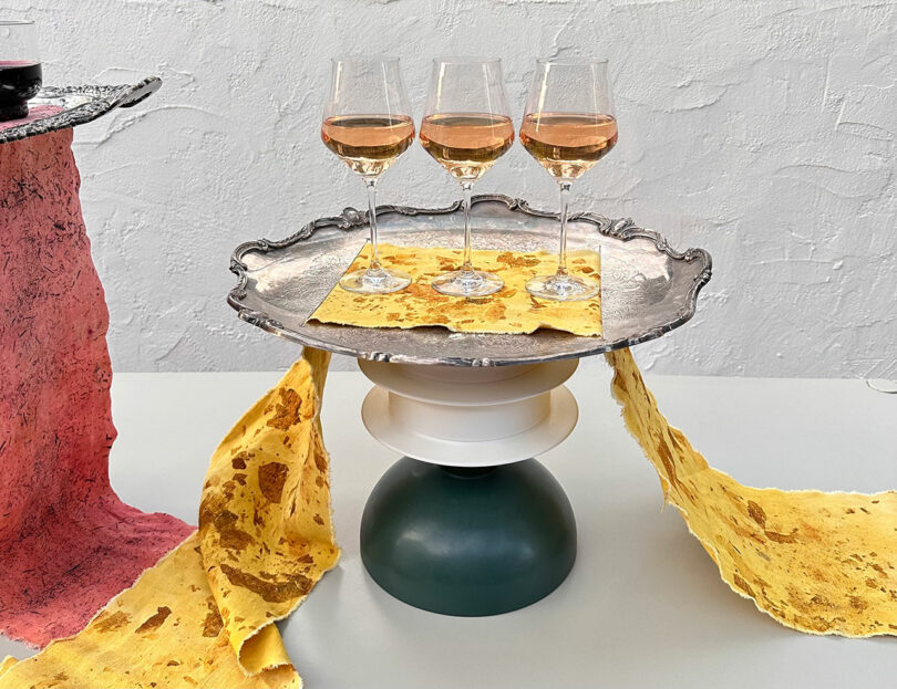 Three glasses of rosé wine on a silver tray, resting on a green and white pedestal, with yellow, textured paper draped over the sides.