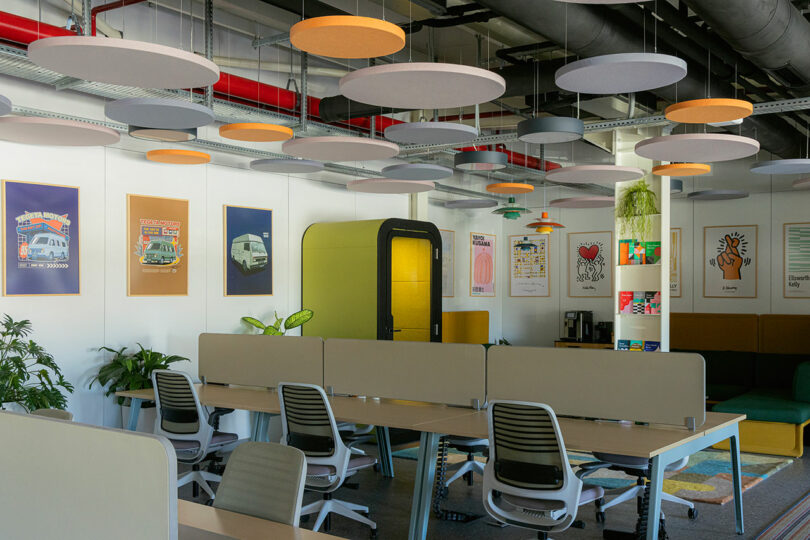 Modern office space with desks, chairs, colorful ceiling panels, wall art, and plants. Cozy seating area with a coffee machine in the background.