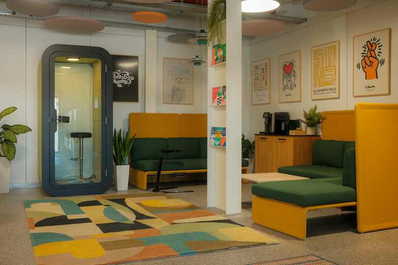 Modern office lounge with mustard and green seating, a phone booth, colorful rug, plants, and framed artwork on walls. Coffee station and decorative ceiling panels complete the decor.