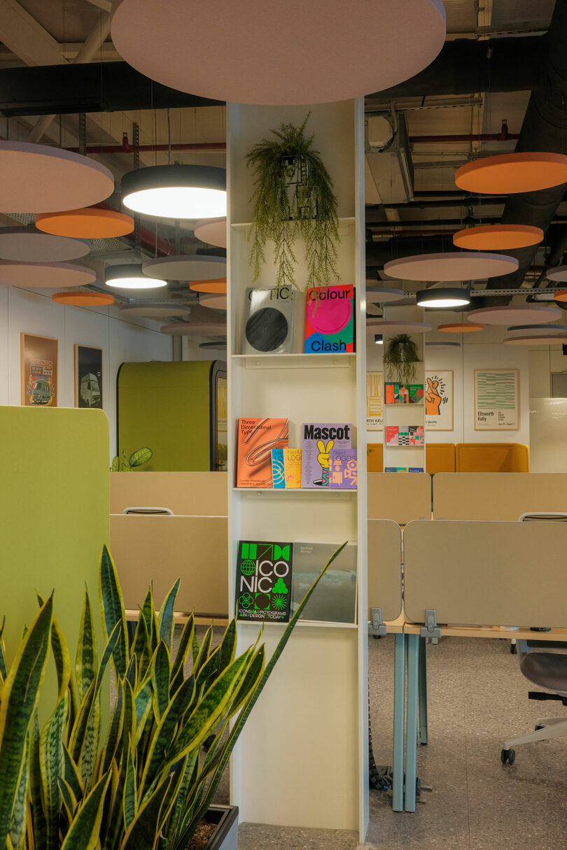 Office interior with plants and colorful books on a white shelf. Modern design with round ceiling panels and partitioned desks.