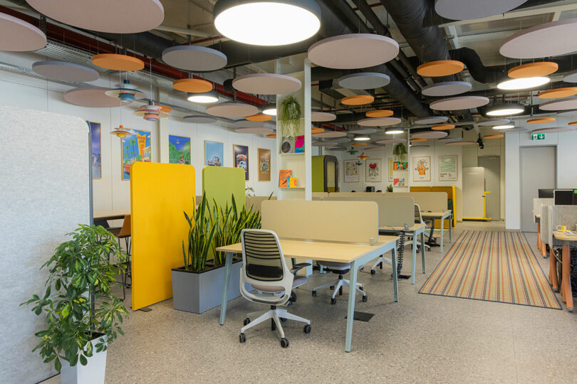 Modern open-plan office space with desks, chairs, plants, and colorful acoustic panels on the ceiling and walls.