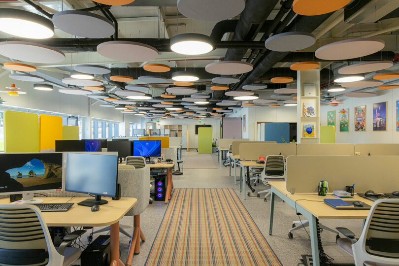 Modern open-plan office with multiple desks, computers, ergonomic chairs, and colorful ceiling acoustics. Bright lighting and partition walls enhance the workspace.